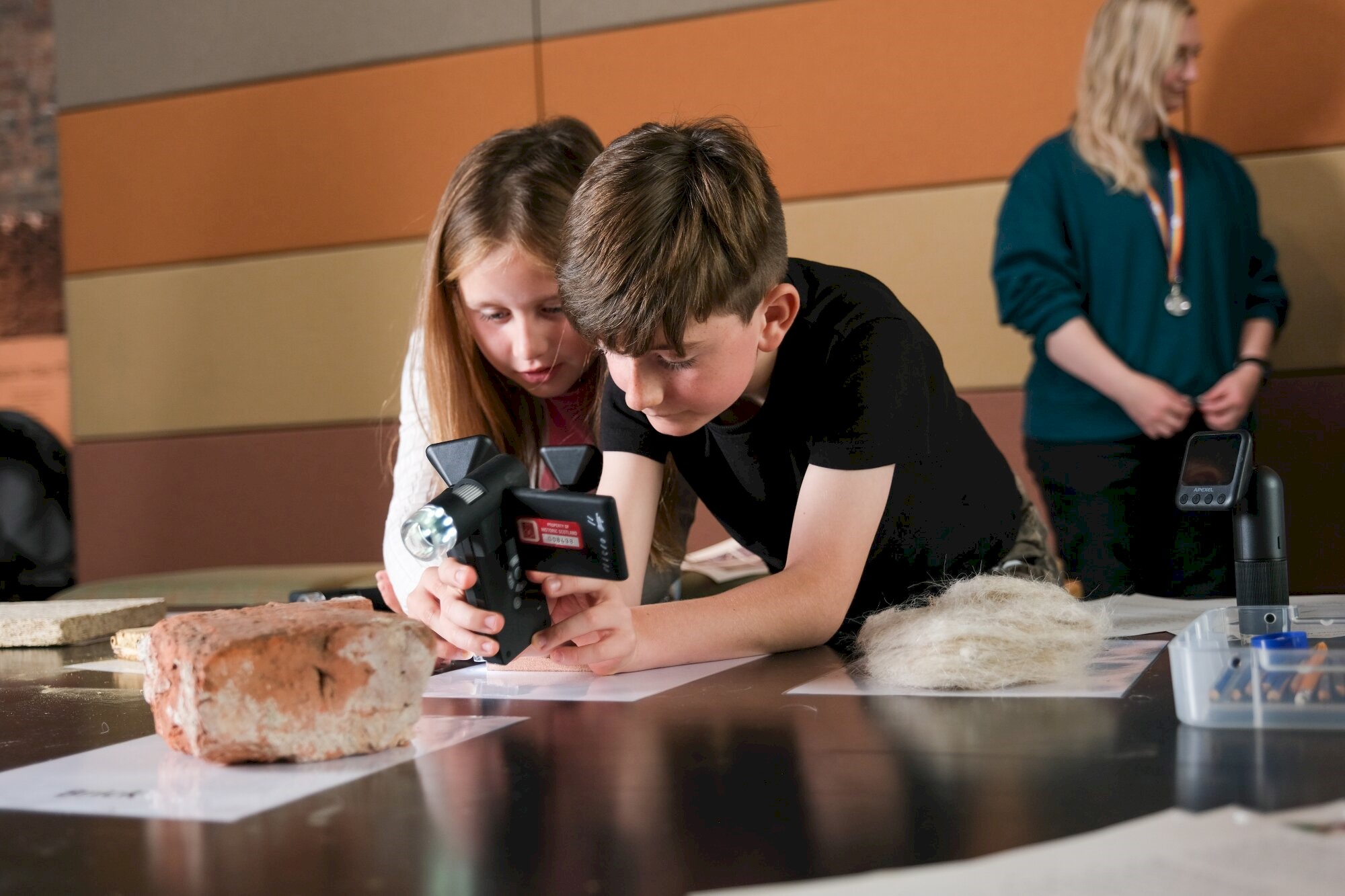 Two young people leaning on a table are working together to point a camera-style piece of equipment at a stone.