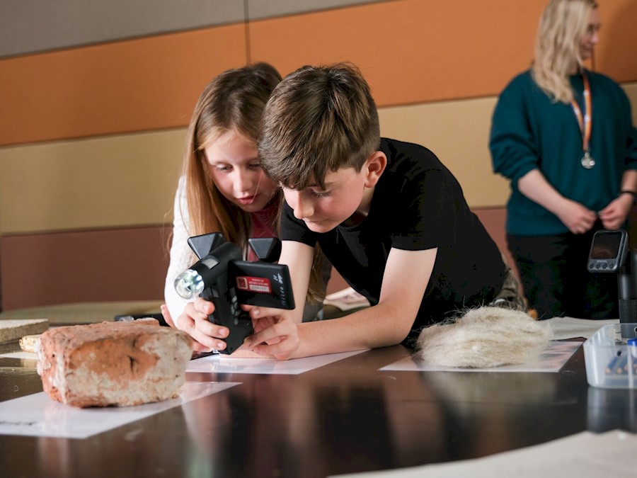 Two young people leaning on a table are working together to point a camera-style piece of equipment at a stone.