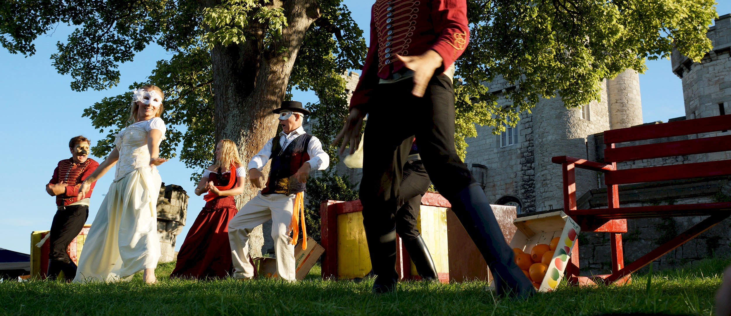 Actors on an outdoor stage performing under a large tree and blue skies
