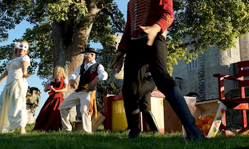 Actors on an outdoor stage performing under a large tree and blue skies