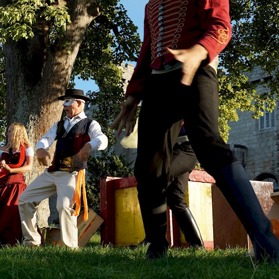 Actors on an outdoor stage performing under a large tree and blue skies