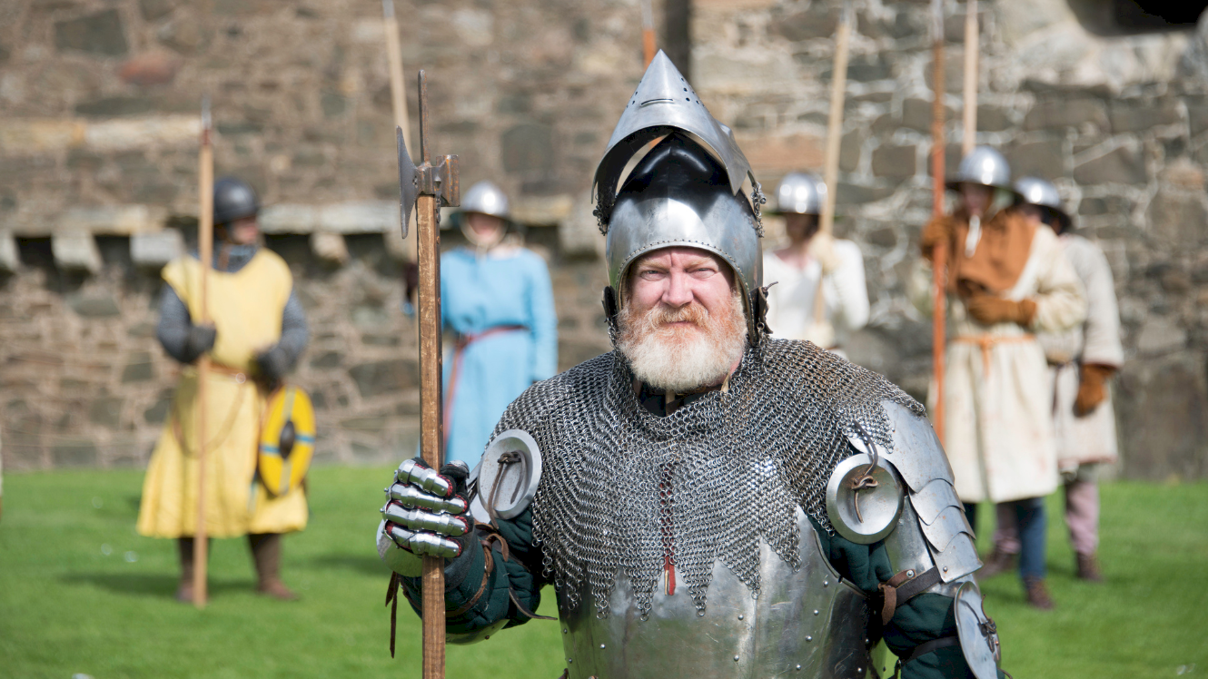 A stern looking costumed performer in chainmail and wearing a helmet, with other fellow costumed performers in the background 