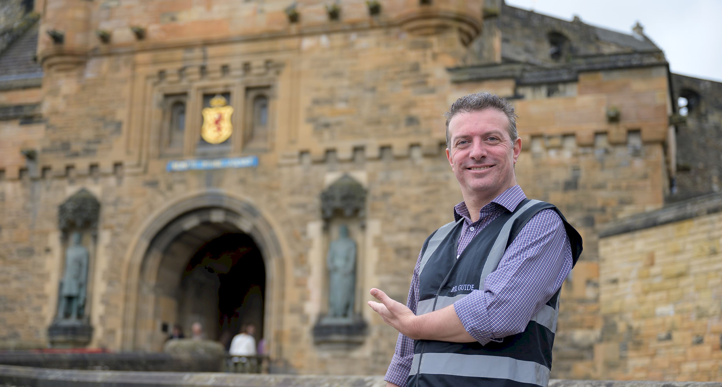 BSL guide Gordon Hay smiling and pointing towards the entrance of Edinburgh Castle