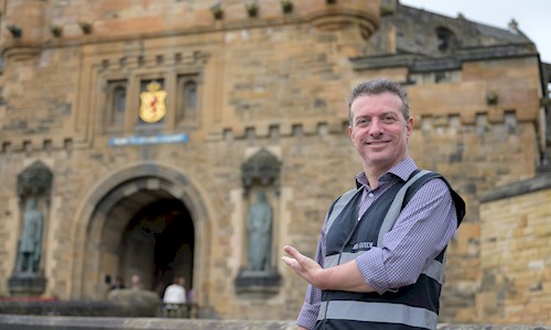 BSL guide Gordon Hay smiling and pointing towards the entrance of Edinburgh Castle