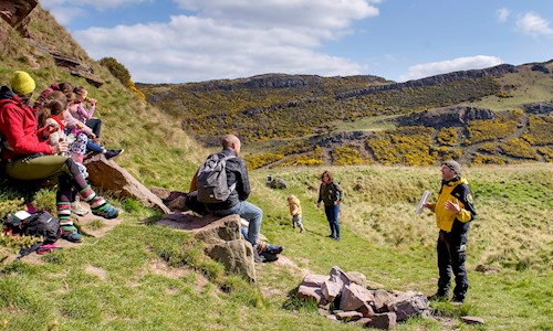 A group of people sitting down listing to a park ranger in Holyrood Park