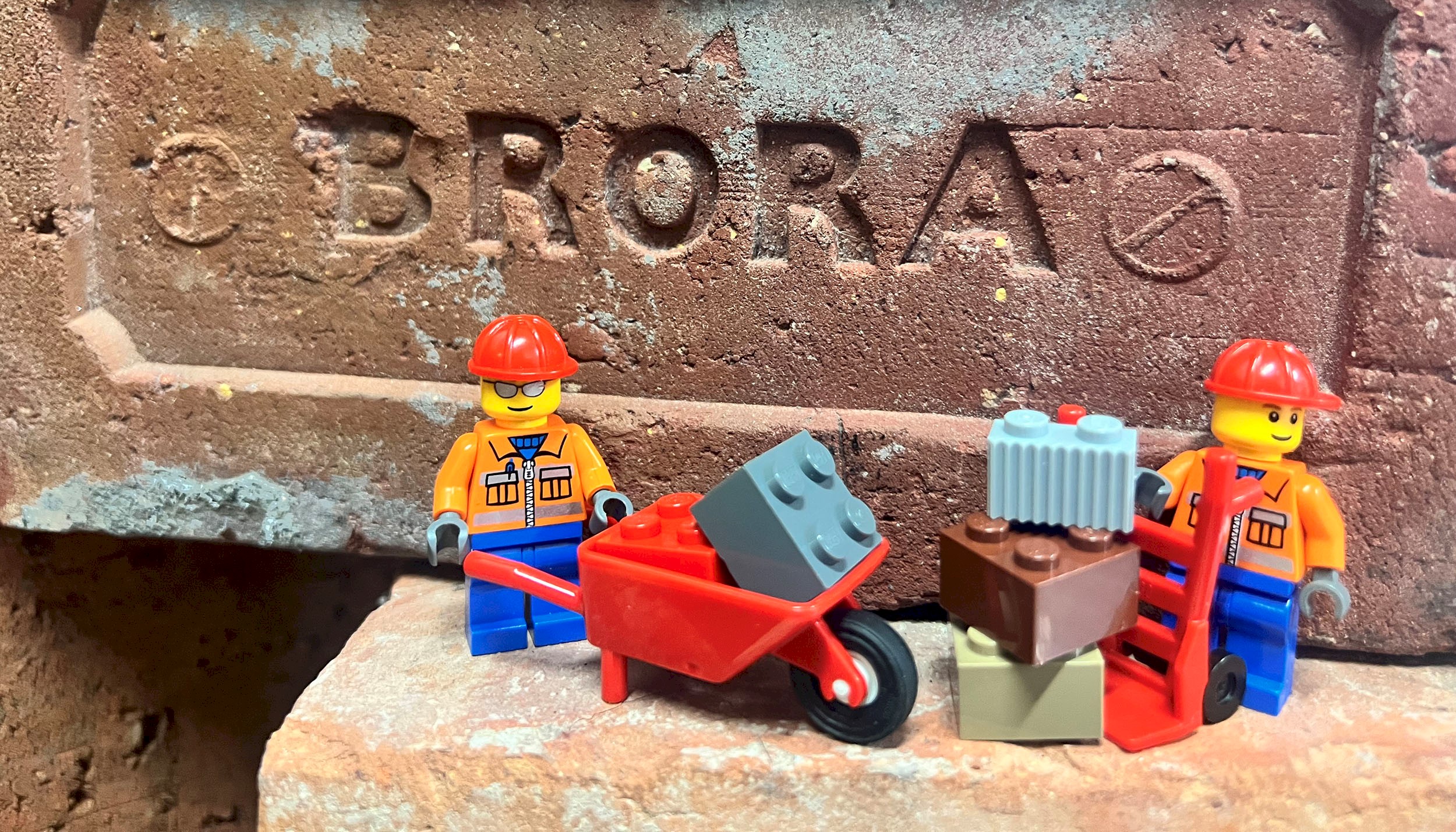 Two LEGO™ figures wearing hard hats, one with a wheelbarrow the other a hand truck. There is a stone brick with the word 'BRORA' carved in