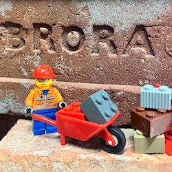Two LEGO™ figures wearing hard hats, one with a wheelbarrow the other a hand truck. There is a stone brick with the word 'BRORA' carved in