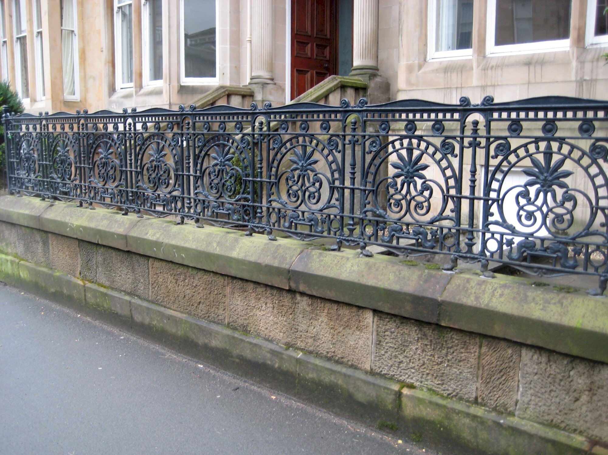 A set of ornate black iron railings outside a row of tenement buildings. The railings have a central circular theme with a large leaf.