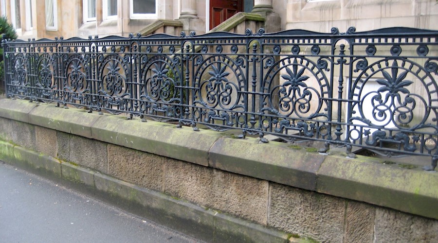 A set of ornate black iron railings outside a row of tenement buildings. The railings have a central circular theme with a large leaf.