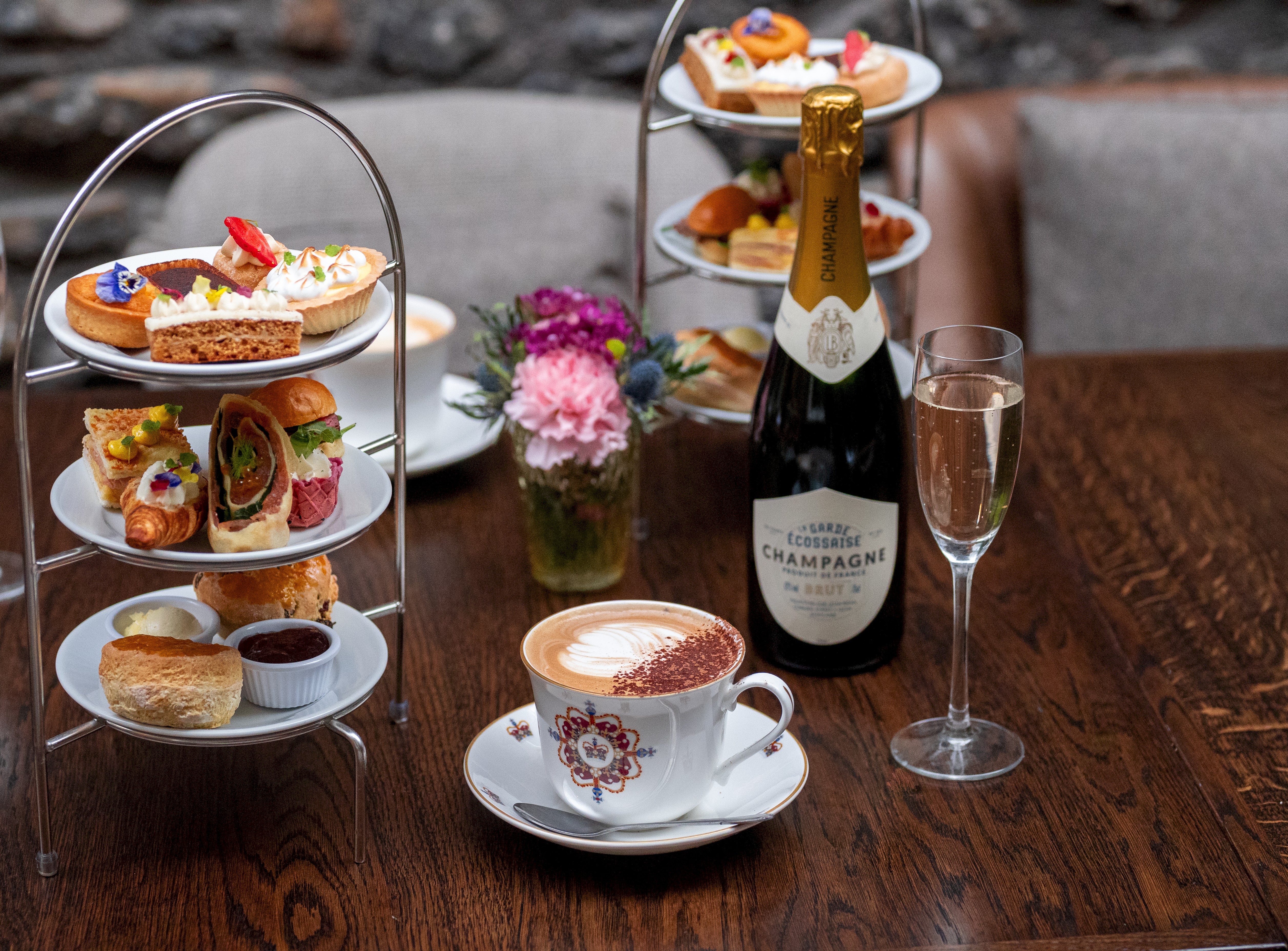 Two tiered cake stands on a table filled with delicious looking cakes and sandwiches, with champagne and coffee on the table too.