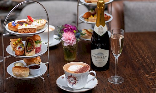 Two tiered cake stands on a table filled with delicious looking cakes and sandwiches, with champagne and coffee on the table too.