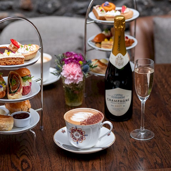 Two tiered cake stands on a table filled with delicious looking cakes and sandwiches, with champagne and coffee on the table too.