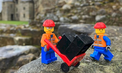 Two LEGO figures in front of Aberdour Castle, one wheeling a trolley with cases on it