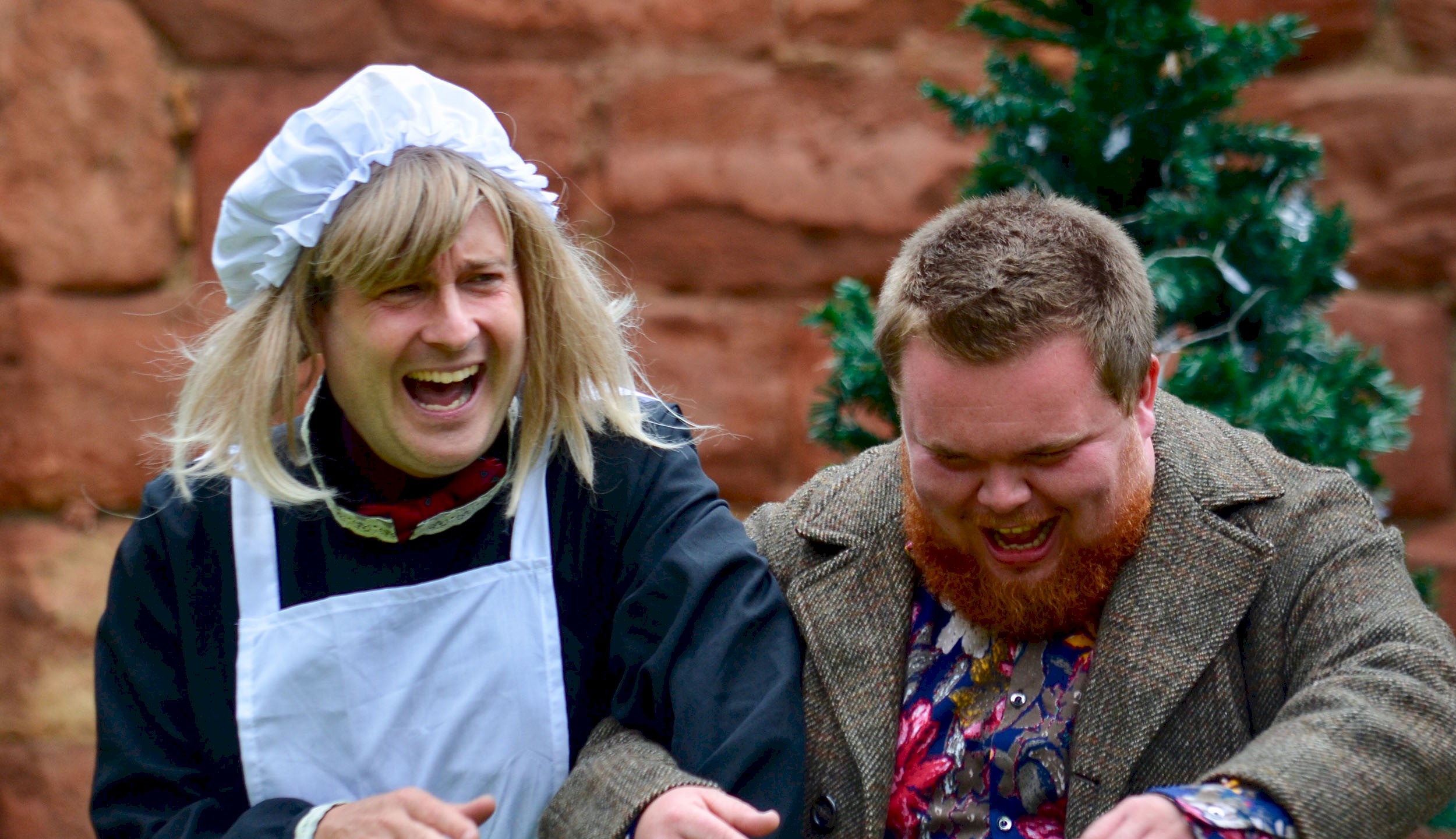 A man wearing a wig dressed as a maid and linked arms with a man with a beard and both laughing heartily