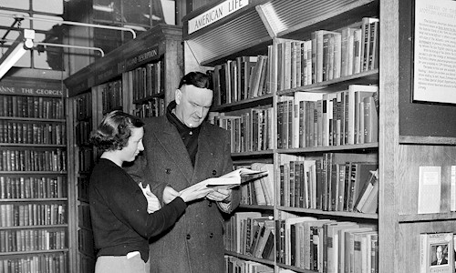 A black and white photo of a woman and man in a library looking at a book