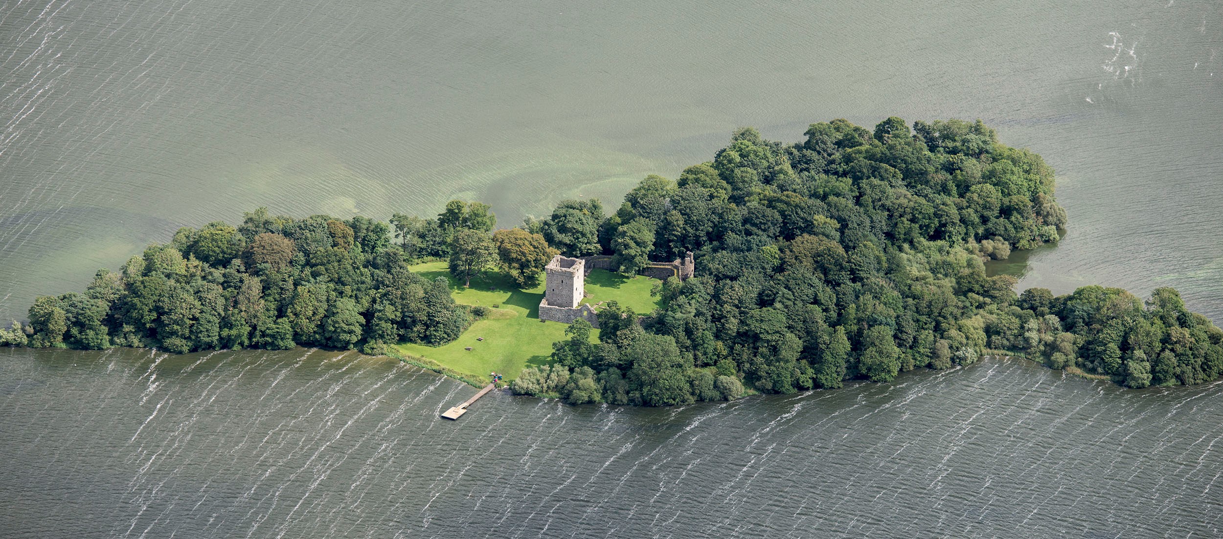 An aerial view of a castle on an island with lots of trees, a pier and surrounded by an expanse of water
