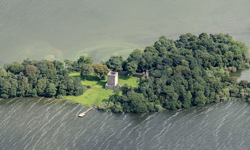 An aerial view of a castle on an island with lots of trees, a pier and surrounded by an expanse of water