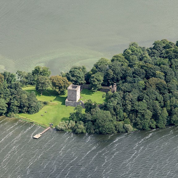 An aerial view of a castle on an island with lots of trees, a pier and surrounded by an expanse of water