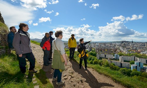 a group of people in walking clothes stand on Arthur's Seat looking out over Edinburgh