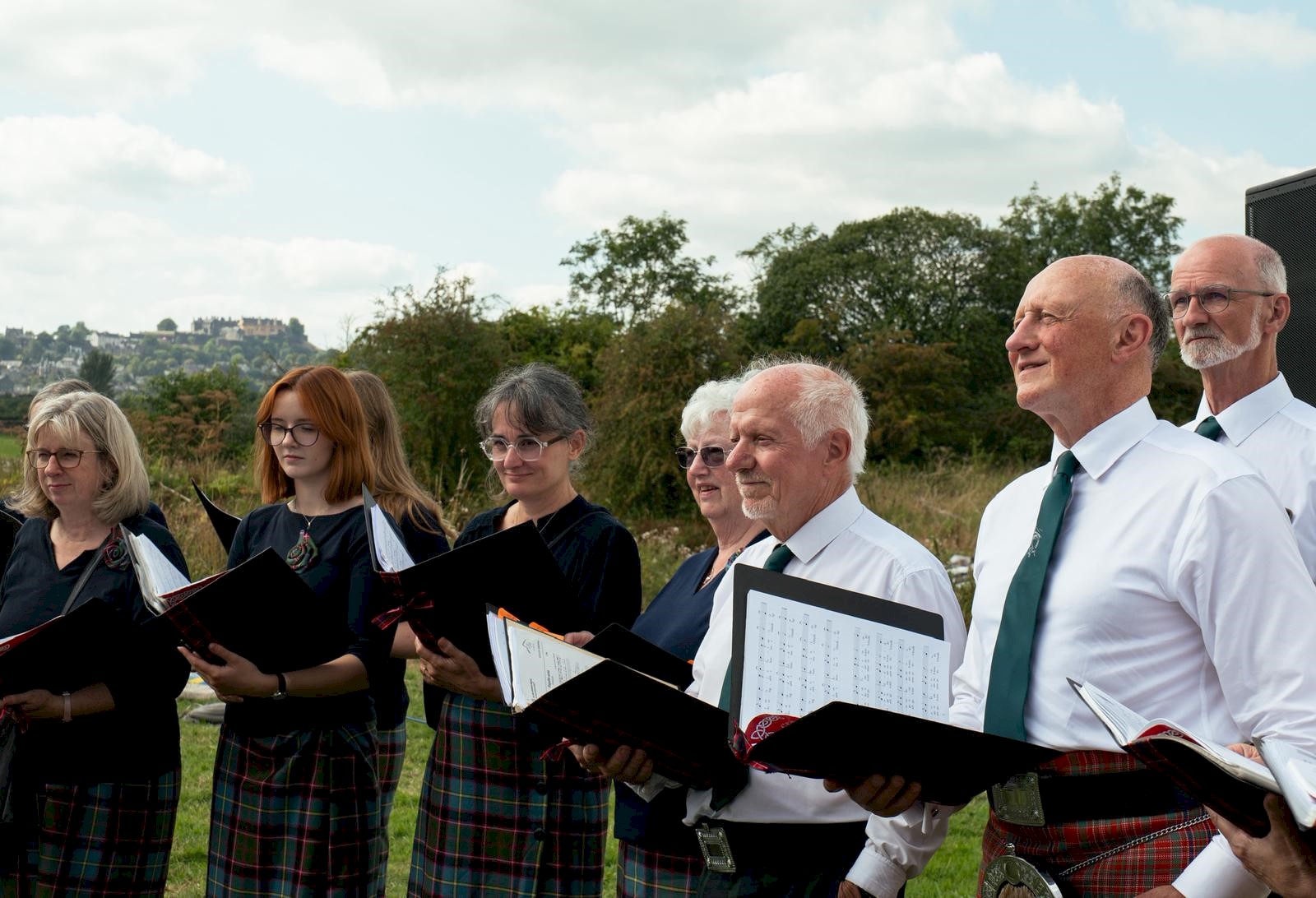 A choir outdoors singing with lots of greenery behind them