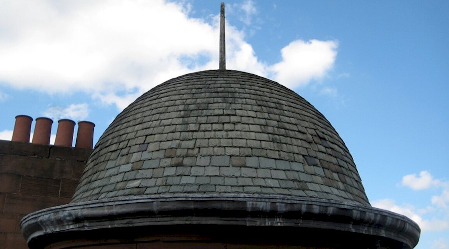 A bell shaped tiled roof of a turreted building, with chimneys in the background. The roof has a spike on the top and the sky is blue with scattered clouds.