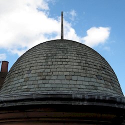 A bell shaped tiled roof of a turreted building, with chimneys in the background. The roof has a spike on the top and the sky is blue with scattered clouds.
