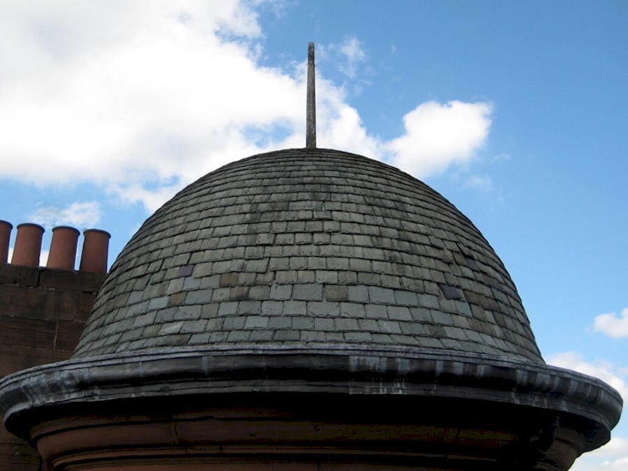 A bell shaped tiled roof of a turreted building, with chimneys in the background. The roof has a spike on the top and the sky is blue with scattered clouds.