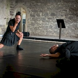 Two performers on a stage, one person is laying on the floor and the other is gasping.