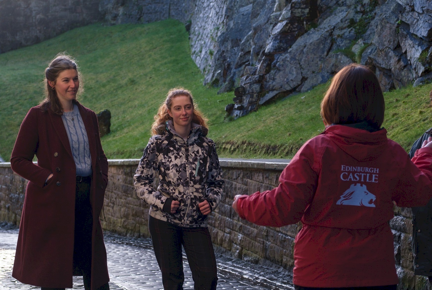 A group of visitors stands on a cobbled pathway beside the stone walls and rocky embankment of Edinburgh Castle. In the foreground, a guide wearing a red jacket with “Edinburgh Castle” printed on the back gestures while addressing the group.