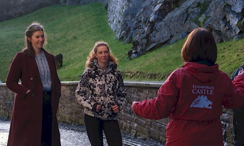 A group of visitors stands on a cobbled pathway beside the stone walls and rocky embankment of Edinburgh Castle. In the foreground, a guide wearing a red jacket with “Edinburgh Castle” printed on the back gestures while addressing the group.