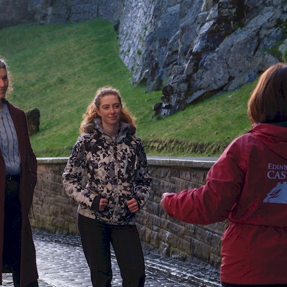 A group of visitors stands on a cobbled pathway beside the stone walls and rocky embankment of Edinburgh Castle. In the foreground, a guide wearing a red jacket with “Edinburgh Castle” printed on the back gestures while addressing the group.