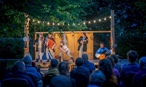 Actors on a lit outdoor stage playing musical instruments and a seated crowd watching on