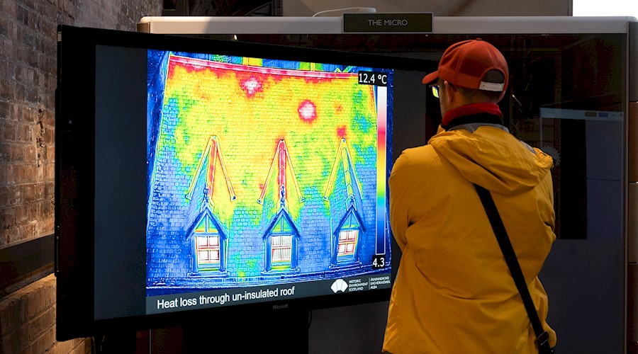 A person wearing a jacket and cap, looking at a large screen which has an image of a house on it that has been taken with a thermal camera showing different colours where there are cold and warm areas