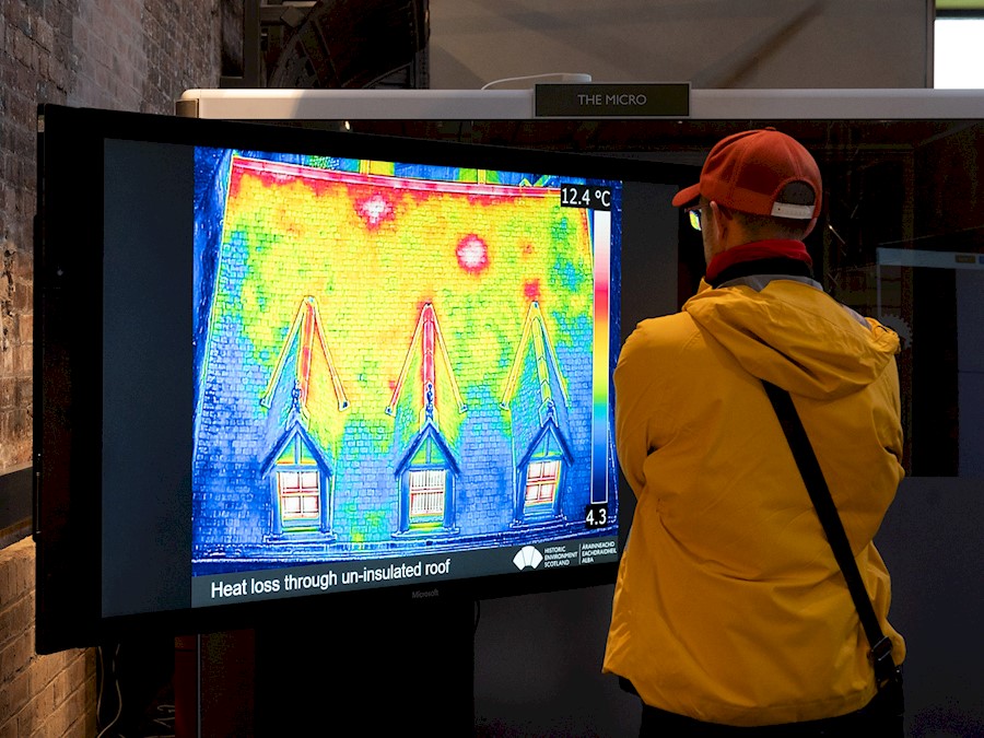 A person wearing a jacket and cap, looking at a large screen which has an image of a house on it that has been taken with a thermal camera showing different colours where there are cold and warm areas