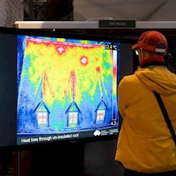 A person wearing a jacket and cap, looking at a large screen which has an image of a house on it that has been taken with a thermal camera showing different colours where there are cold and warm areas