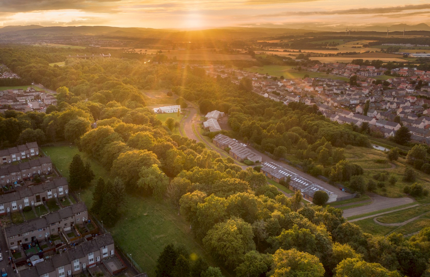 Image is an aerial photograph of a landscape of trees and houses. The houses are at either side, in the centre of the landscape is a large stretch of trees running in a straight line to the horizon. The sun is setting over the hills in the distance.
