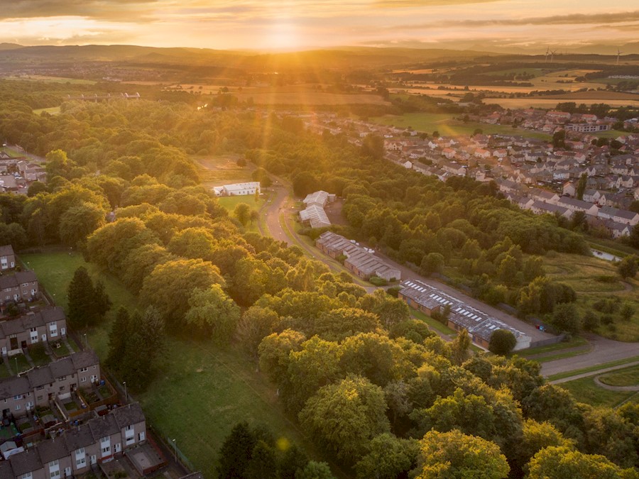 Image is an aerial photograph of a landscape of trees and houses. The houses are at either side, in the centre of the landscape is a large stretch of trees running in a straight line to the horizon. The sun is setting over the hills in the distance.