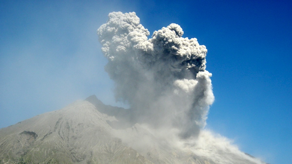 A volcano erupting with grey/white smoke and a very blue sky