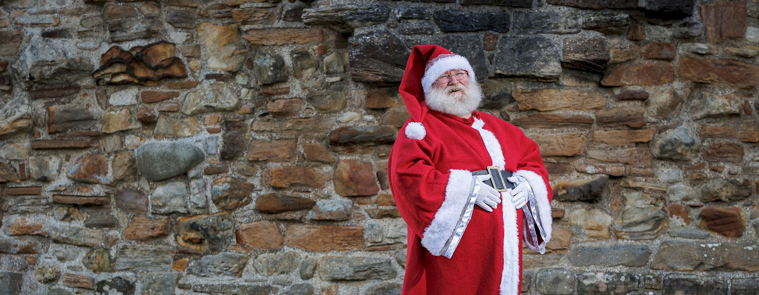 A man standing in front of a high brick wall dressed as Santa Claus