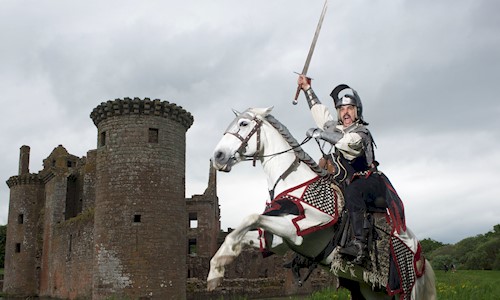 Costumed performer dressed as a Knight on a horse wielding a knife with Caerlaverock Castle in the background