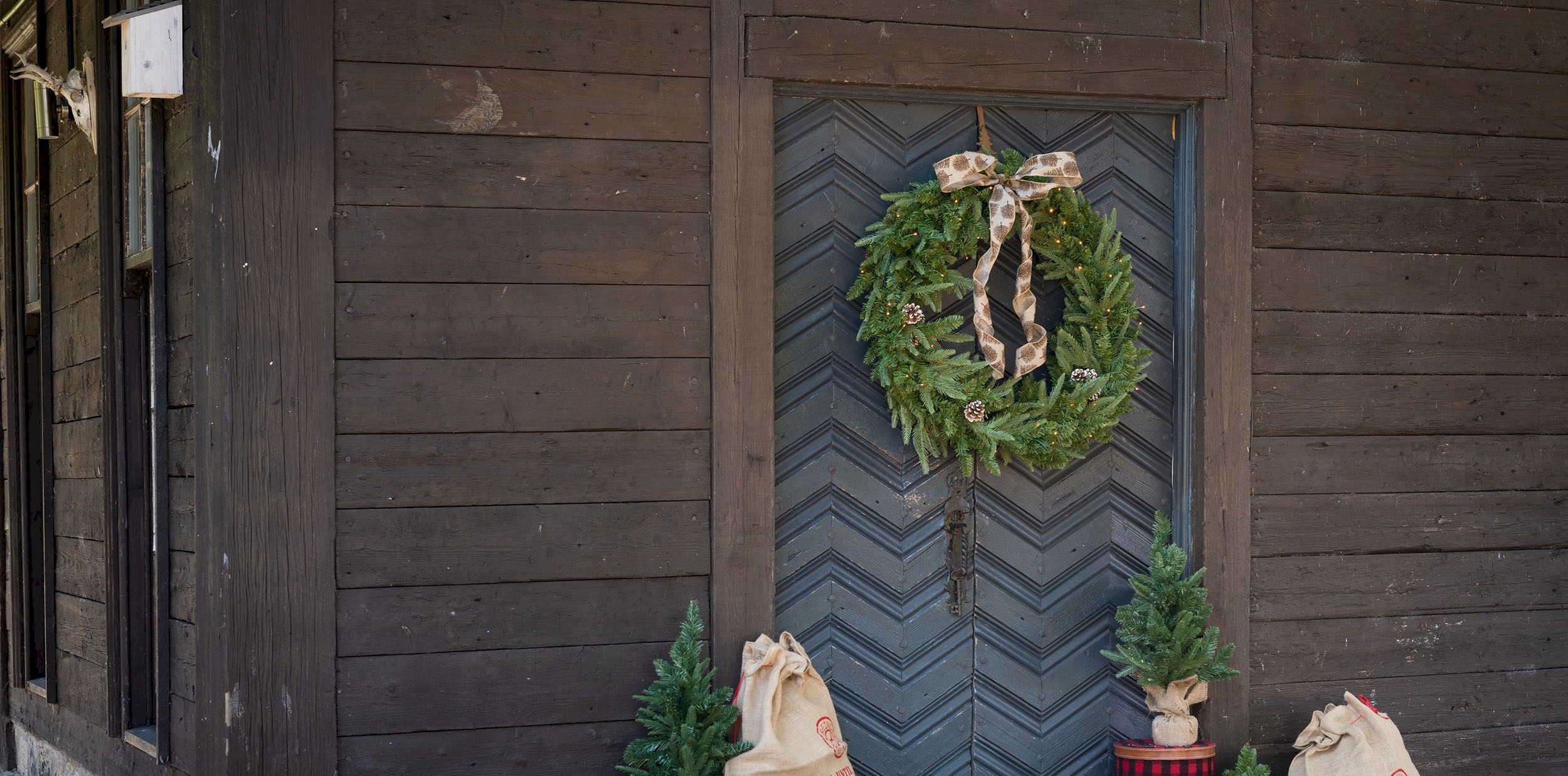 A wooden building with a wreath on the door and some small Christmas trees and present sacks on the ground