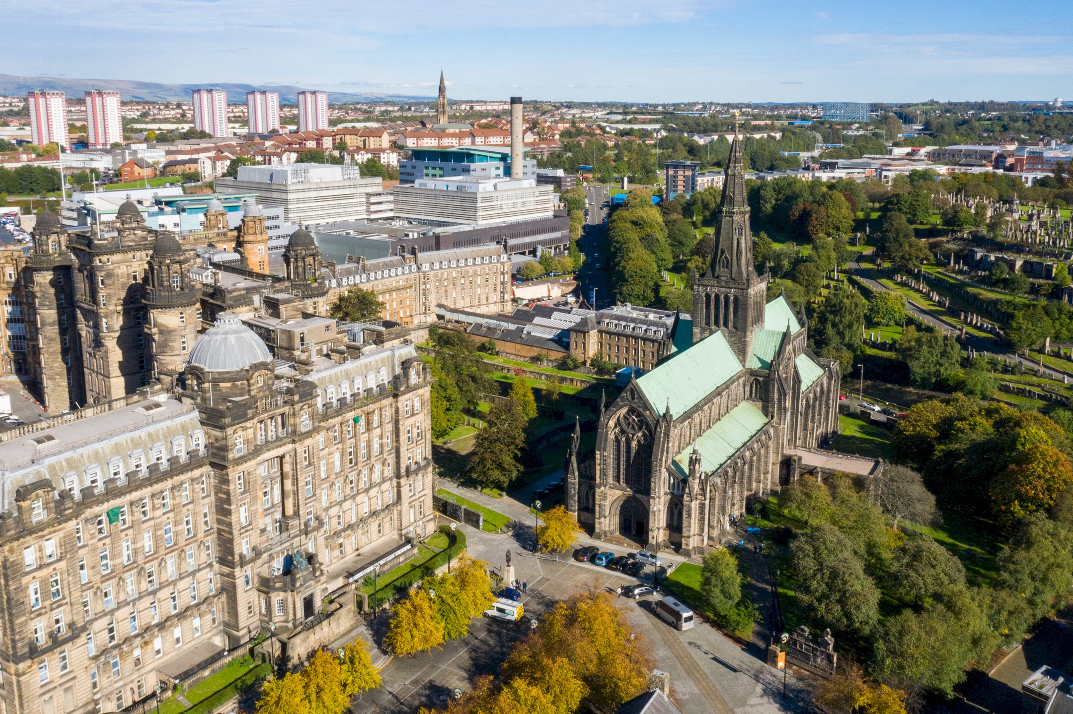 Aerial view Glasgow city centre showing large stone civic buildings and a Gothic cathedral with a tall spire and green copper roof, surrounded by trees and parkland.