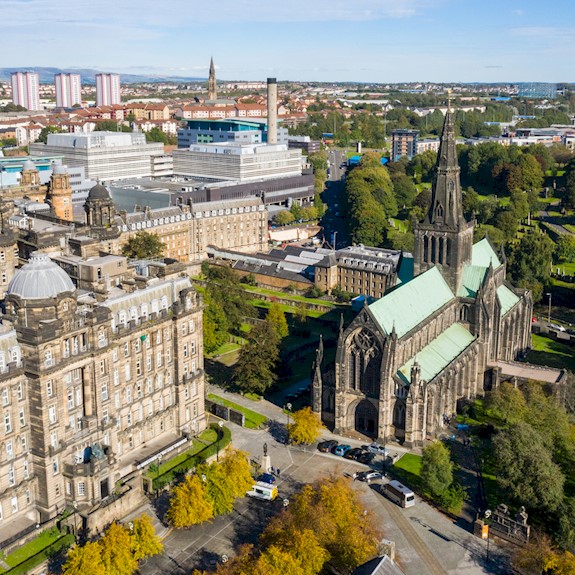 Aerial view Glasgow city centre showing large stone civic buildings and a Gothic cathedral with a tall spire and green copper roof, surrounded by trees and parkland.