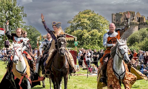 5 people in costume on horses with a crowd watching on and a castle in the background