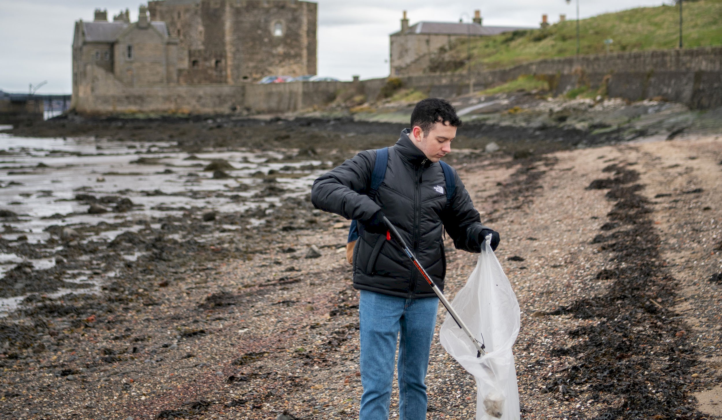 A man on a shingle beach picking up litter with a castle in the background