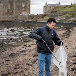 A man on a shingle beach picking up litter with a castle in the background