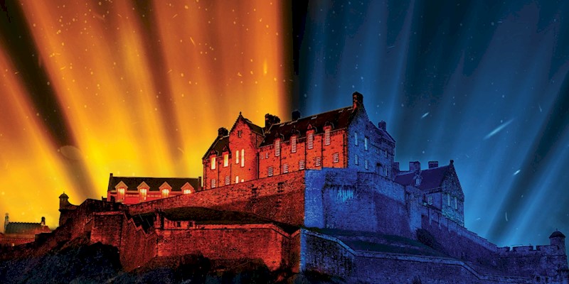 Edinburgh Castle illuminated in vibrant colors for the Castle of Light: Fire and Ice campaign. The left side of the castle glows with warm orange and red lights, while the right side is highlighted in cool blue tones, creating a dramatic fire and ice effect against the night sky.