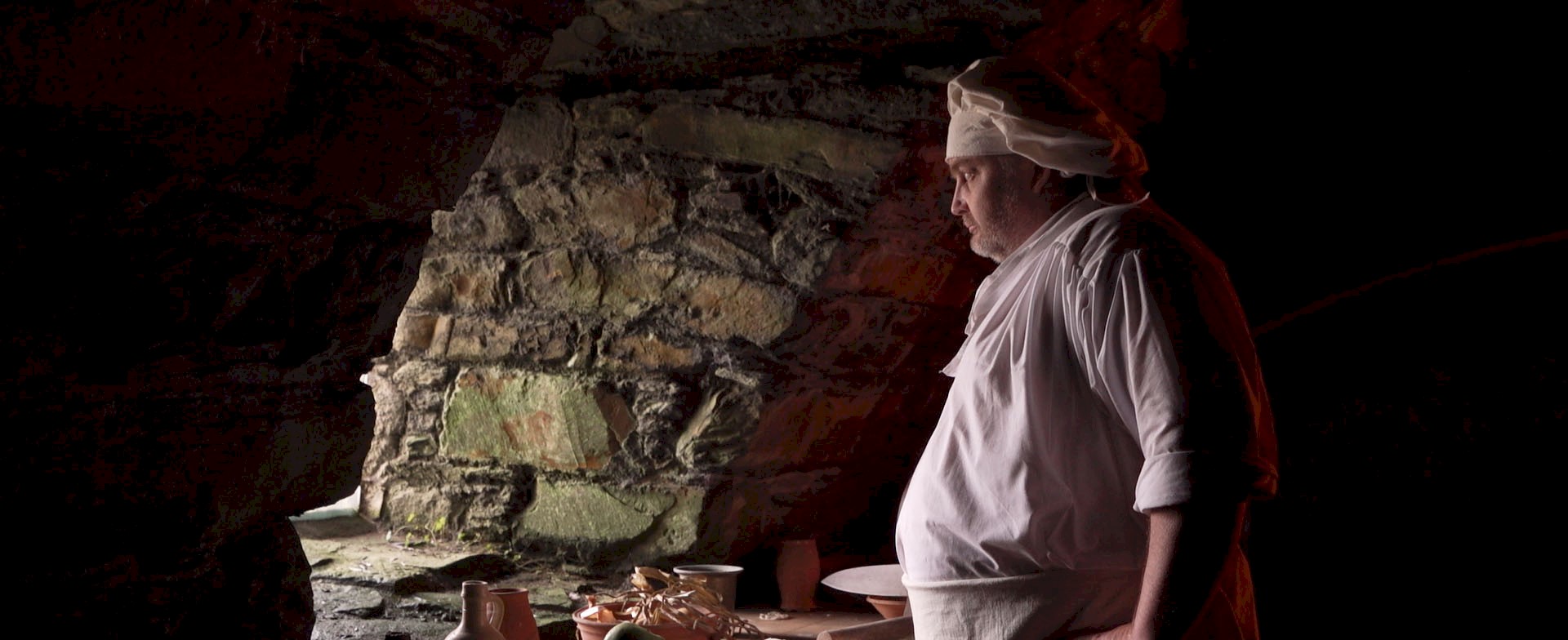 A man in historical chef whites standing over a chopping board with onions and apples, looking out of a window of a stone building