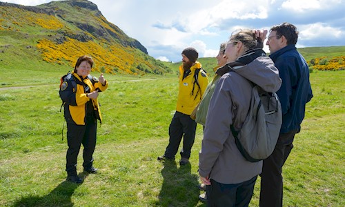 People dressed in outdoor clothes, standing below Arthur's Seat on a sunny day.