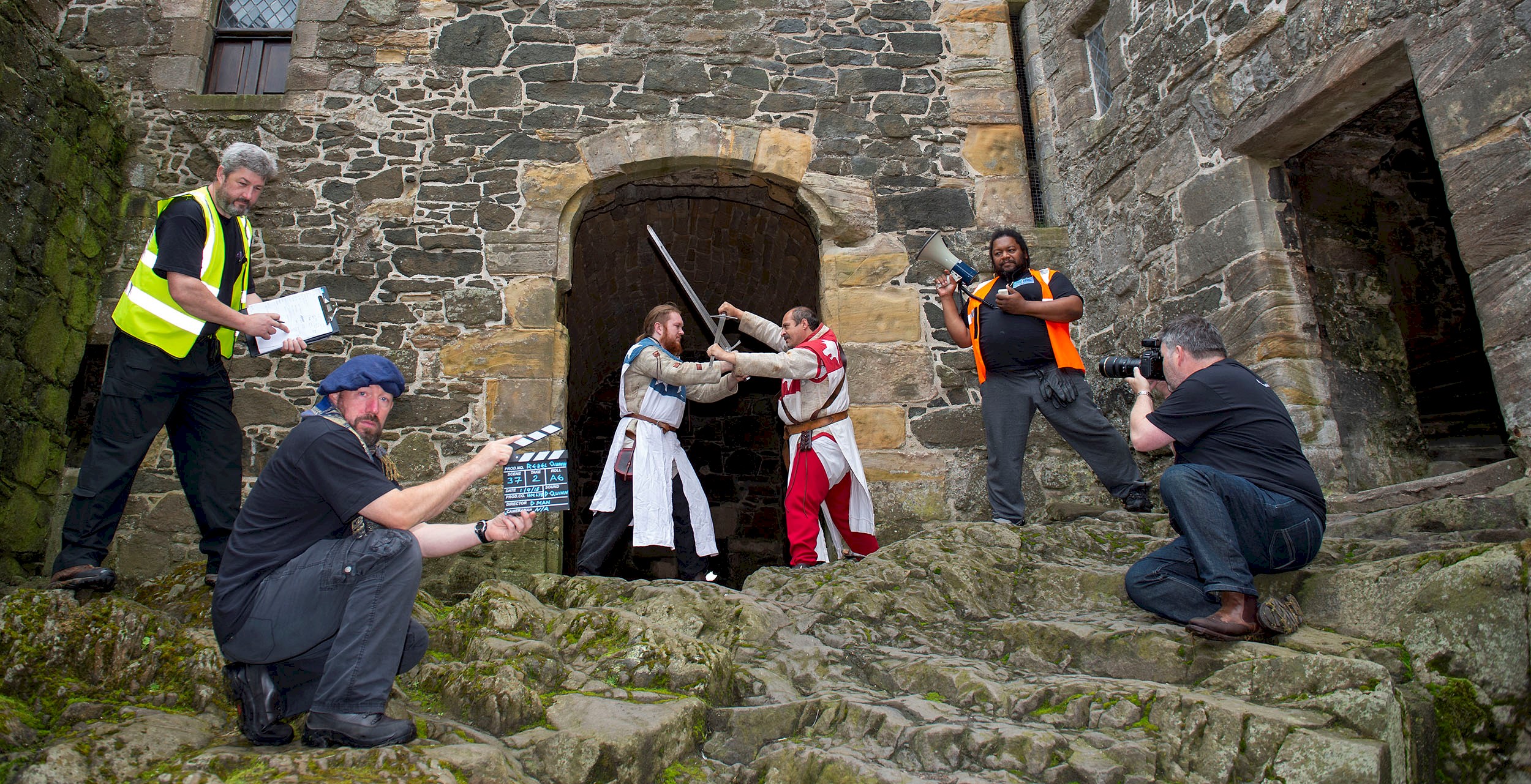 Two re-enactors in the grounds of a castle battling with swords and a crew of 4 with cameras, videos and clapper board